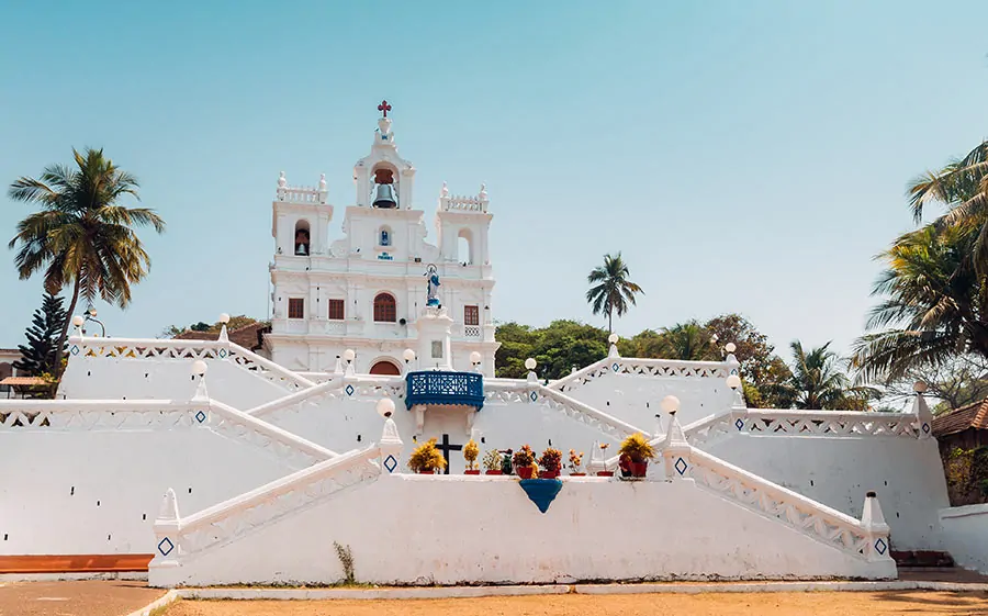 Biały Kościół Niepokalanego Poczęcia Najświętszej Marii Panny w Goa, Indie, z charakterystycznymi schodami i palmami w tle pod jasnoniebieskim niebem