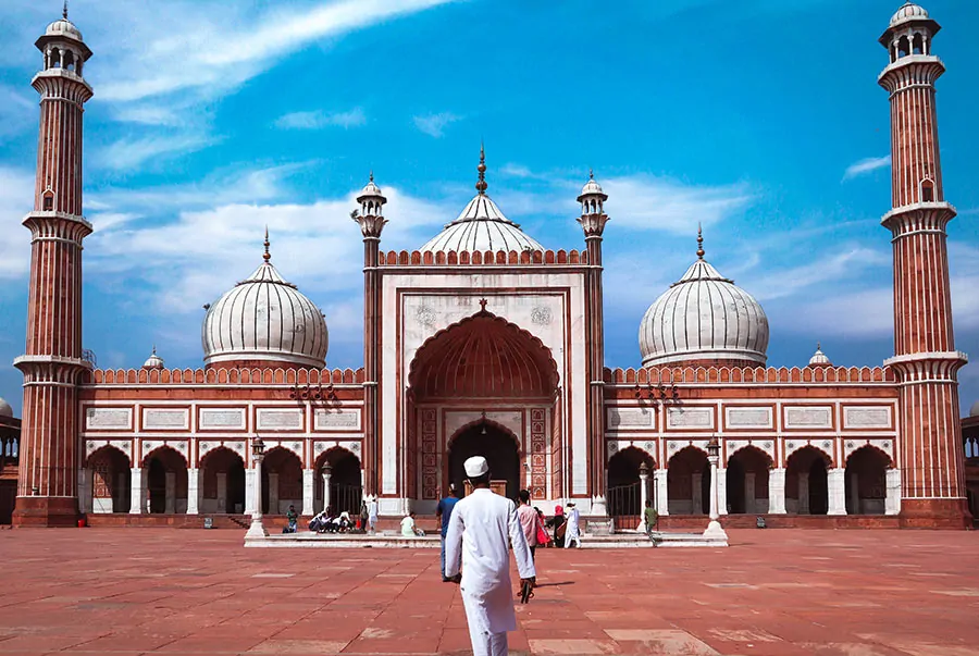 Muzułmanin w białym stroju idzie w kierunku meczetu Jama Masjid w Delhi, Indie, z wieżami minaretów i kopułami w tle. Islam w Indiach.