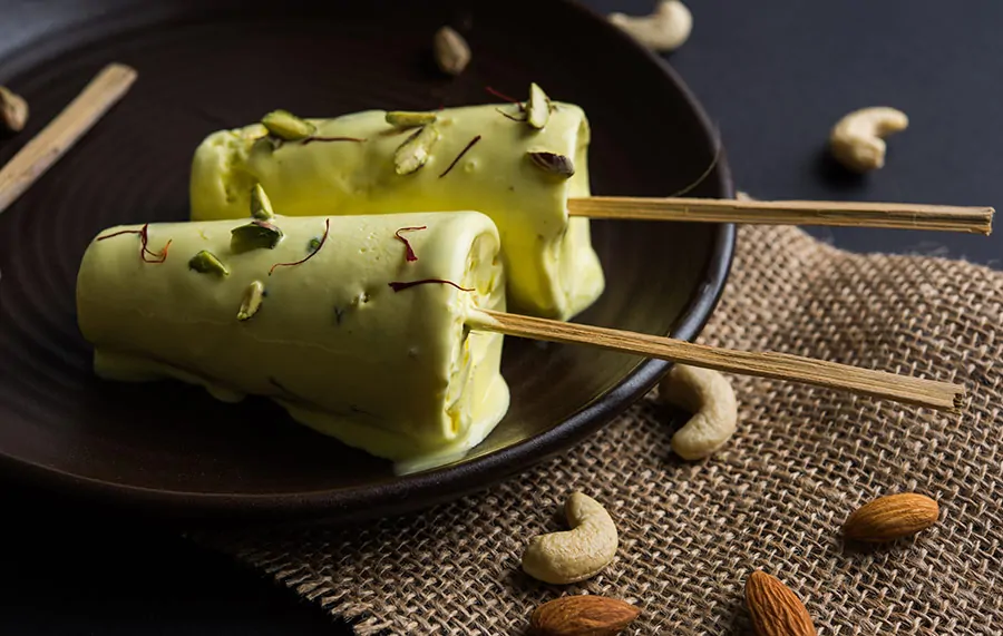 Kulfi, kremowe indyjskie lody na patyku, posypane pistacjami i szafranem, podane na talerzu z orzechami nerkowca i migdałami.