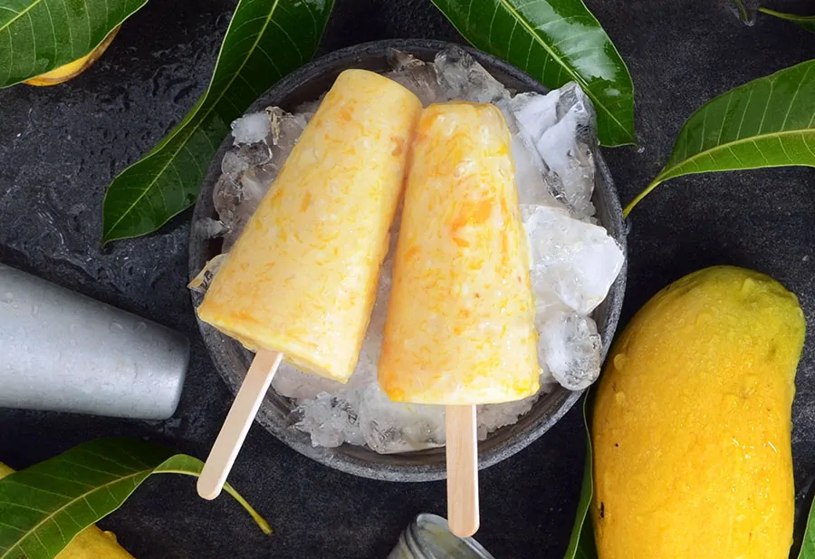 Mango kulfi, indyjskie lody na patyku z kawałkami mango, podane na lodzie, otoczone owocami mango.