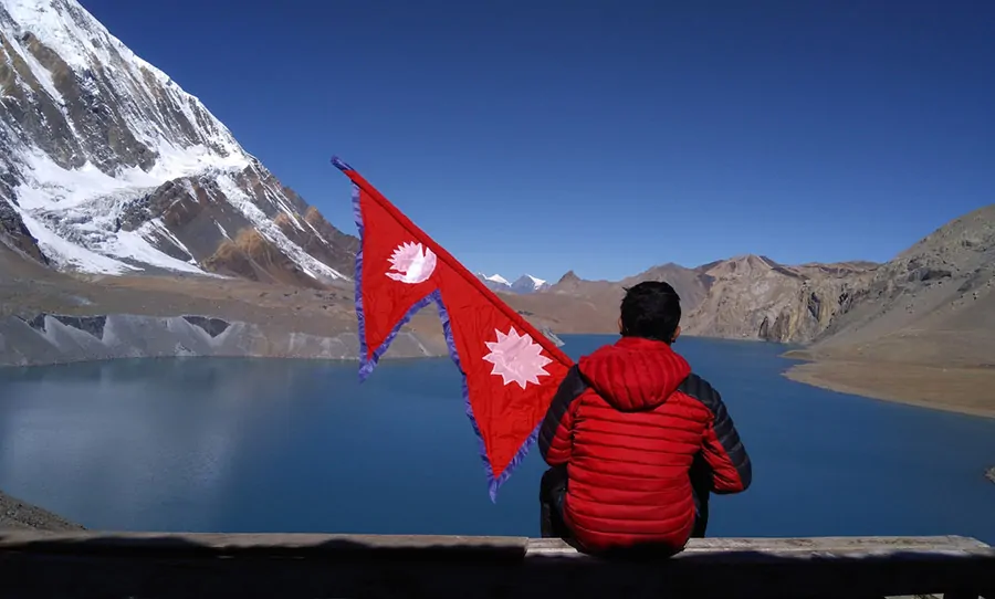 Osoba w czerwonej kurtce siedzi przed spokojnym górskim jeziorem, trzymając w górze flagę Nepalu, ze szczytami Himalajów w tle