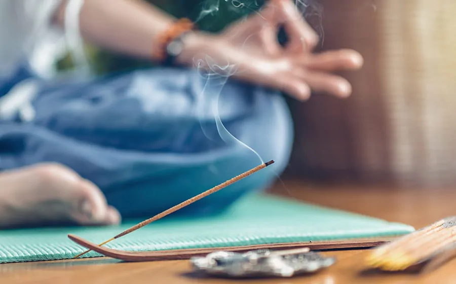 Indyjskie kadzidełko na drewnianej podstawce z delikatnym dymem na tle osoby praktykującej medytację.